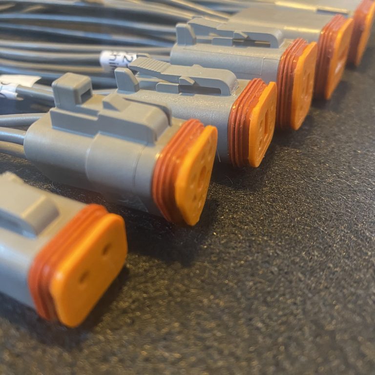 A lineup of Voltforge Deutsch DT pigtail assemblies on a dark workshop surface, grey wire with heatshrink cavity idents visible, orange wedgelocks seated on the connector faces.